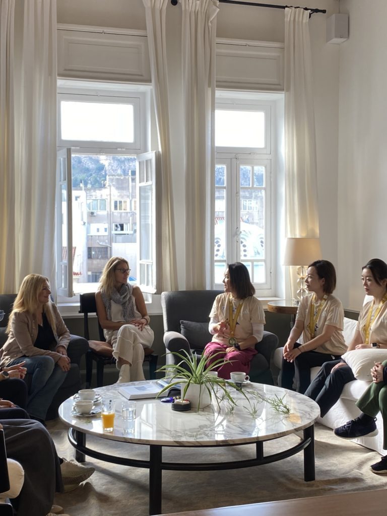 The image depicts a group of five women engaged in a casual meeting in an elegantly furnished room with large windows draped with long white curtains. The room is bathed in natural light, suggesting a daytime setting. They are seated around a round marble coffee table with a plant centerpiece, various beverages, and some papers, indicating an informal discussion or a social gathering. The women appear relaxed and are dressed in comfortable, casual business attire, with lanyards around their necks, which could suggest they are at a conference or professional event. The outside view through the windows shows a cityscape with buildings and a glimpse of a hill in the distance, possibly indicating an urban location.