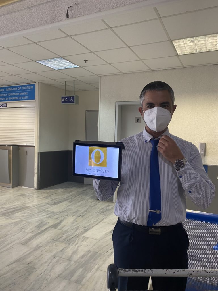 A man in a professional attire consisting of a light blue, striped shirt with a dark blue tie and trousers stands in an indoor setting, possibly an office or a travel-related public area. He is wearing a white mask over his mouth and nose, adhering to health and safety protocols. He is holding a digital tablet displaying the logo of "MY ODYSSEY TRAVEL OFFICES" which features a large golden "O" on a blue background. The environment includes signage in the background, including one for a "W.C." indicating restroom facilities, and another sign that reads "MINISTRY OF TOURISM OPERATION OF TOURISM OF CRETE," suggesting this may be a location associated with travel or tourism in Crete, Greece.