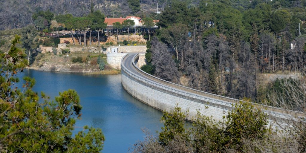 The image displays Marathon Lake in Greece, showcasing a curving dam structure made of concrete, stretching across the lush green landscape. The dam curves gracefully from the left to the right side of the frame, bordering the serene blue waters of the lake. Behind the dam, there is a dense patch of trees on a hillside, with a few structures partially visible amongst the foliage. Wind turbines stand on the distant hills, signifying a blend of natural beauty and renewable energy. The sky is clear, suggesting a calm and bright day, ideal for visiting this historic and scenic location.