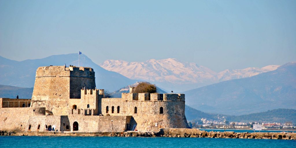 The image features the Bourtzi Castle, a Venetian fortress located in the middle of the harbor of Nafplio, Greece. The castle stands prominently on a small islet, connected to the mainland by a pier visible in the foreground. The fortress is made of stone, showing signs of weathering, and has a round central tower with additional battlements. A Greek flag is hoisted on top of the structure, and visitors can be seen walking around its base. In the background, majestic snow-capped mountains rise under a clear blue sky, creating a striking contrast against the calm turquoise waters of the sea. The scene captures a blend of Greece's rich historical architecture with its natural beauty.