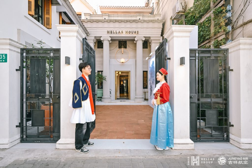 Two individuals in traditional Greek attire standing outside the entrance of Hellas House.