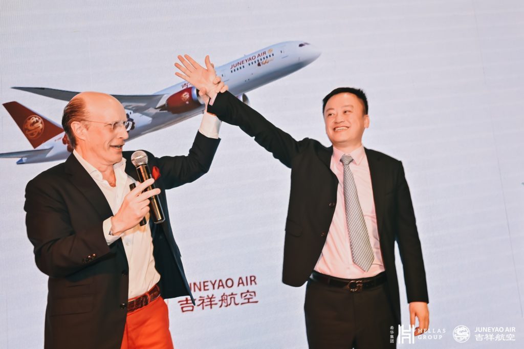 Two men on stage sharing a high-five, one holding a microphone, with a backdrop displaying an airplane and the logos of Hellas Group and Juneyao Air.