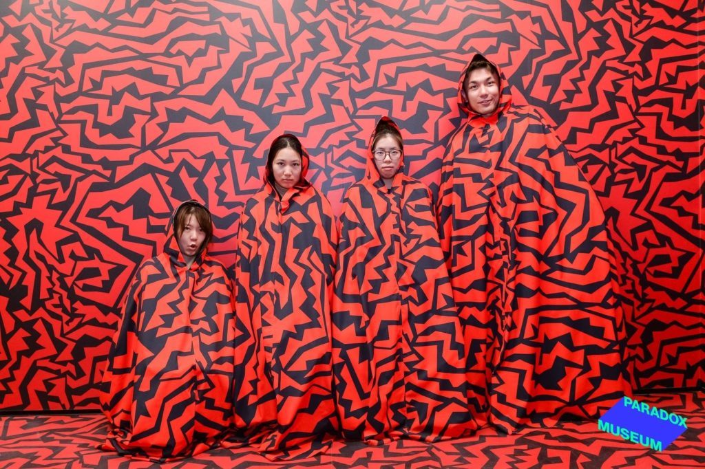 Four people standing against a red and black maze-patterned backdrop, wearing capes that camouflage them into the background, at the Paradox Museum.