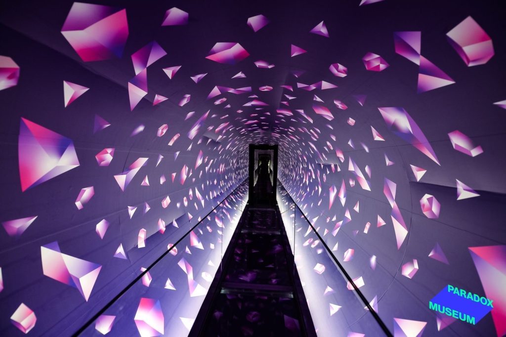 A vibrant tunnel with illuminated pink and purple geometric shapes on the walls, leading to an exit, inside the Paradox Museum.