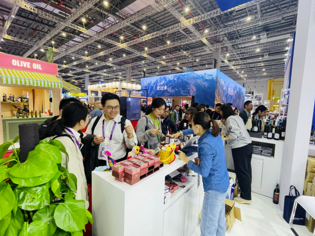 Visitors sampling Greek products at the “Best of Greece” booth during CIIE in Shanghai, featuring olive oil and wine displays and a busy trade show atmosphere.